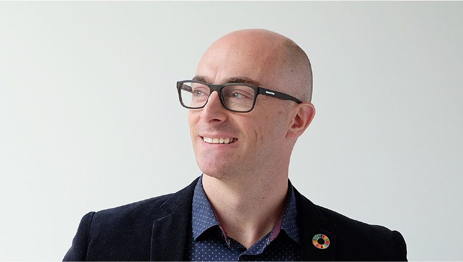 A man in a suit looks to his right. He is wearing an SDG pin.